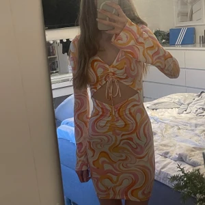 Färgglad klänning från H&M - Säljer en färgglad klänning från H&M med ett mönster i rosa, orange och vitt. Klänningen har långa ärmar och en snygg knytdetalj framtill. Klänningen har några fläckar som förmodligen går bort i tvätten😊