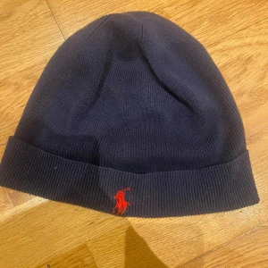 Mörkblå mössa från Polo Ralph Lauren - Mörkblå ribbstickad mössa från Polo Ralph Lauren med klassisk röd broderad logga framtill. Enkel och stilren design som passar till många olika outfits. Tillverkad i mjukt material för extra komfort.