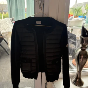 Svart cardigan jacka från Moncler - Säljer en svart cardigan jacka från Moncler med quiltade paneler framtill och mjuka stickade ärmar. Jackan har rund halsringning, dragkedja och Moncler-logga på ärmen. Perfekt för lager-på-lager och snygg till många outfits. Storlek M men passa från M-Xs 