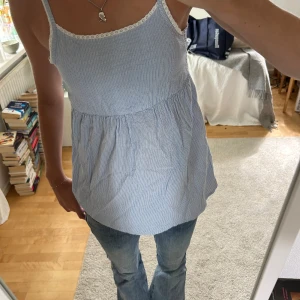 Blå och vit randig linnetopp med spetsdetaljer - Säljer en superfin blå och vit randig linnetopp i storlek S. Toppen har smala axelband och är dekorerad med söt spets längs kanterna. Perfekt för sommaren och kan stylas med jeans eller shorts för en avslappnad look. Toppen är i bra skick och redo för en ny ägare!