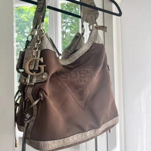Brun axelväska från Guess - Säljer en snygg brun axelväska från Guess med coola detaljer i silver. Väskan har ett stort Guess-logotyp på framsidan och är gjord i ett slitstarkt material. Den har också en praktisk dragkedja och stiliga kedjor som ger den en extra touch. Perfekt för både vardag och fest! Pris går att diskutera 💕