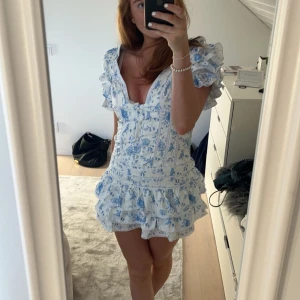 Blommig kortklänning med volanger - Säljer en superfin blommig kortklänning i ljusblått och vitt. Den har en v-ringad design med volanger på ärmarna och kjolen, vilket ger en romantisk känsla. Perfekt för sommarens fester eller en dag på stan!