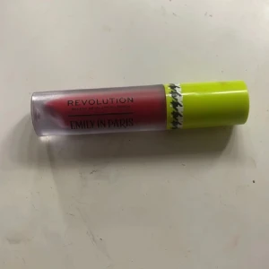 Rött läppglans från Revolution - Säljer ett snyggt röd blush från Revolution med temat 'Emily in Paris'. Det har en cool limegrön kork med ett svartvitt mönster. Perfekt för att ge dina kinder en pop av färg och glans!