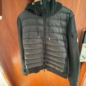 Svart pufferjacka från Moncler - Säljer en snygg svart pufferjacka från Moncler. Jackan har en quiltad design med dragkedja och en huva med snörning. Den är perfekt för kyligare dagar och har en Moncler-logga på ärmen. Jackan är i bra skick och redo för nya äventyr!