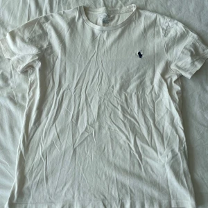 Vit t-shirt från Polo Ralph Lauren - Säljer en klassisk vit t-shirt från Polo Ralph Lauren i storlek M. Den har en liten broderad logga på bröstet och är i en slim fit passform. Perfekt för en stilren och enkel look. T-shirten är i bomull och har korta ärmar. (Se sista bild med ett litet hål, syns bara om man kollar noga). Frågor? Skriv!
