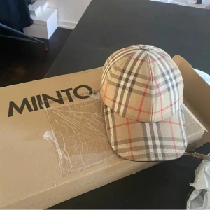 Beige keps från Burberry - Säljer en snygg beige keps från Burberry med det klassiska rutmönstret i svart, rött och grått. Kepsen är i storlek M och är perfekt för både vår och sommar. Den är helt ny och kommer i originalförpackning. Perfekt för att ge din outfit en lyxig touch! Perfekt skick och knappt använd.