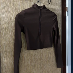 Brun ribbad croppad tröja med dragkedja - Säljer en snygg brun croppad tröja med ribbad textur och dragkedja framtill. Perfekt för en stilren och modern look. Tröjan har långa ärmar och en tight passform som framhäver figuren. Passar perfekt till både vardag och fest!