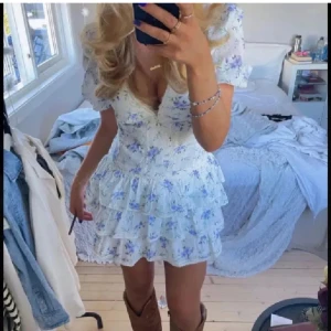 Blommig kortklänning med volangdetaljer - Säljer en superfin blommig kortklänning med volangdetaljer. Klänningen har en v-ringad design och korta ärmar, perfekt för sommarens fester. Den är i en ljus färg med blå och lila blommor, vilket ger en romantisk känsla. Materialet känns lätt och luftigt, perfekt för varma dagar💕 Lånad bild!