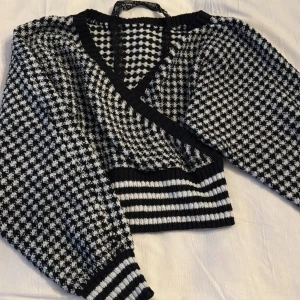 Svartvit stickad omlottopp - Säljer en snygg svartvit stickad omlottopp med ett rutigt mönster. Toppen har långa ärmar och ribbade muddar vid ärmslut och nederkant. Perfekt för en trendig look!
