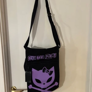Svart axelväska med lila tryck - Cool svart axelväska med justerbar rem och ett unikt lila tryck av en katt med rosett och korsade benknotor. Perfekt för att ge en edgy touch till din outfit.