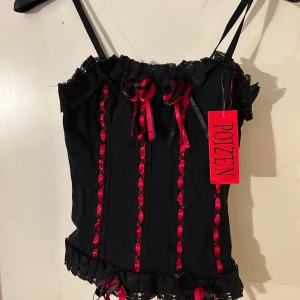 Svart korsettopp med röda detaljer från Poizen - Snygg svart korsettopp från Poizen med röda snörningsdetaljer och spetskant. Axelbandslös design med dekorativa rosetter framtill. Perfekt för en unik och edgy stil.