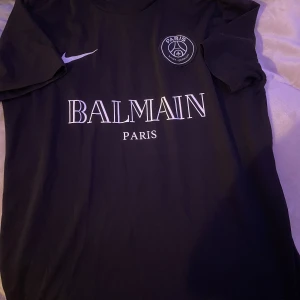 Svart PSG fotbollströja från Nike - Snygg svart PSG fotbollströja med Balmain Paris-logga på bröstet. Tröjan har korta ärmar och en klassisk rund halsringning. 