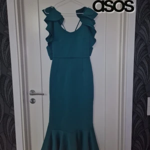 Grön Klänning med Volanger - Elegant mörkgrön klänning från ASOS – storlek 40 – 499 kr    Säljer denna stilfulla mörkgröna klänning från ASOS med vackra volangdetaljer. Perfekt för bröllop, fest, bal eller andra speciella tillfällen!    Detaljer: ✔ Märke: ASOS   ✔ Färg: Djup mörkgrön   ✔ Modell:** Figurnära passform med volangärmar och volangdetalj vid fållen   ✔ Rygg:bSnyggt skuren med korsade band för en elegant look   ✔ Material: Bekvämt och följsamt tyg som sitter fint på kroppen   ✔ Storlek: 40    Skick: ✔ Mycket fint 
