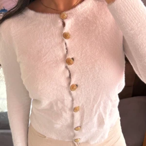 Vit kofta med guldknappar - Säljer en elegant vit kofta med unika guldknappar. Den är långärmad och har en mjuk, fluffig textur som ger en lyxig känsla. Perfekt för att lyfta vilken outfit som helst.