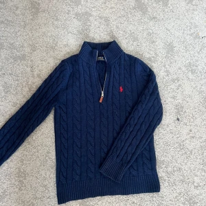 Mörkblå stickad tröja från Polo Ralph Lauren - Säljer en snygg mörkblå stickad tröja från Polo Ralph Lauren med dragkedja och röd logga på bröstet. Tröjan har ett klassiskt flätmönster och ribbade muddar vid ärmslut och nederkant. Perfekt för kyliga dagar!