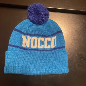 Ljusblå Nocco mössa - Snygg blå mössa med mörkblå tofs och uppvikt kant. Mössan har NOCCO-logga i vitt och mörkblå ränder. Perfekt för kalla dagar!