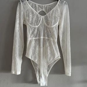 Vit spetsbody - Elegant vit spetsbody med långärmad design och öppen rygg. Den har ett vackert mönster och en smickrande passform. Perfekt för att lägga till en touch av elegans till din garderob. Liten i storleken. 
