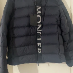  Moncler Ume - Snygg svart dunjacka från Moncler med dragkedja och stor logga framtill. Jackan har en hög krage och avtagbar huva, perfekt för kyliga dagar. Den quiltade designen ger en stilren look. Pris kan diskuteras 