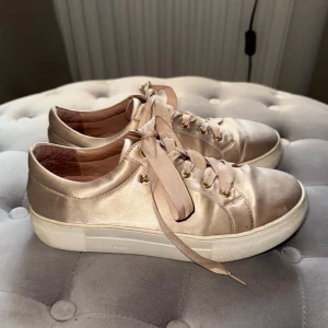 Beige sneakers med satinfinish - Snygga beige sneakers med satinfinish och breda snören. Skorna har guldiga detaljer vid snörningen och är perfekta för en stilren look. Bara använt ett få tal gånger eftersom jag växte ur dem🩶 Märket är K.cobler