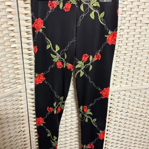 Svarta leggings med rosor - Snygga svarta leggings med ett blommigt mönster av röda rosor och gröna blad. Perfekta för att lägga till en färgklick i din outfit. De har en tight passform och är gjorda i ett stretchigt material för extra komfort.