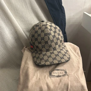 Beige keps från Gucci - Snygg beige keps från Gucci med det klassiska GG-mönstret. Kepsen har en justerbar rem baktill och en röd och grön randdetalj på sidan. Perfekt för att ge din outfit en lyxig touch.