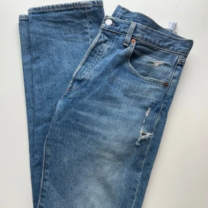 Levis 501 jeans med slitningar - Klassiska Levis 501 jeans i blå denim med coola slitningar på benen och bakfickan. Perfekta för en avslappnad och trendig look. De har en knappgylf och fem fickor. Storlek 30/34
