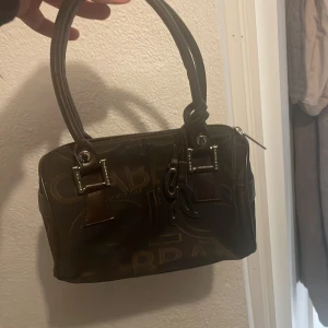 Brun handväska med mönster - Snygg brun handväska med ett unikt mönster och dubbla handtag. Väskan har en dragkedja upptill och detaljer i metall. Perfekt för att bära dina dagliga nödvändigheter med stil.