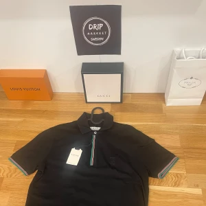 Small Svart pikétröja från Lufian - Snygg svart pikétröja från Lufian med färgglada ränder på kragen och ärmsluten. Tröjan är tillverkad i bomull med en liten del elastan för extra komfort. Perfekt för en stilren look.