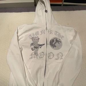 Vit zip hoodie med tryck - Säljer en vit hoodie med dragkedja och ett unikt tryck på framsidan. Texten 'AIM FOR THE MOON' och en tecknad figur pryder tröjan i glittriga detaljer. Perfekt för en avslappnad stil. Inga defekter