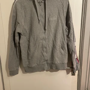 Grå hoodie från Moschino - Snygg grå hoodie från Moschino med dragkedja och huva. Den har långa ärmar med Moschino-logga längs ärmarna och ribbade muddar. Perfekt för en avslappnad stil. Kan gå ner i pris vid snabb affär