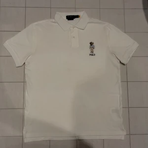 Vit pikétröja från Ralph Lauren - Säljer en stilren vit pikétröja från Ralph Lauren med en broderad björn på bröstet. Tröjan har en klassisk krage och knappar framtill. Perfekt för en avslappnad men ändå elegant look. (Använd endast en gång)