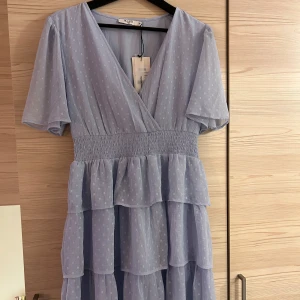 Blå klänning med volanger - Charmig blå klänning med v-ringning och korta ärmar. Klänningen har ett smockat midjeband och flera lager av volanger, 3 lager i kjolen och sedan ärmarna. Helt ny med lapparna kvar, jättegullig klänning som bara inte kommit till användning! 