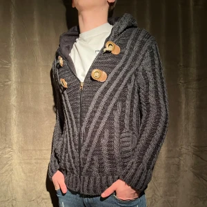 Fodrad Cardigan Jacka - Säljer en mörkgrå stickad cardigan med dragkedja och dekorativa knappar. Tröjan har en mysig huva och ribbade muddar. Storlek L men passar M. Den saknar spännena men det är inget som stör! Perfekt för kyliga dagar. Vid minsta fråga eller fundering hör av er🤝