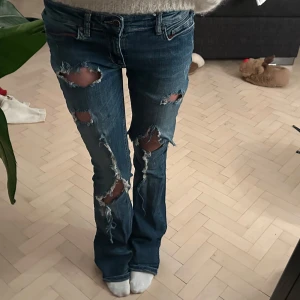 Blå slitna jeans - Säljer ett par blå jeans med slitningar och hål för en cool look. De har en bootcut-stil och är lågmidjade, vilket ger en avslappnad känsla. Perfekta för dig som vill ha en edgy stil.