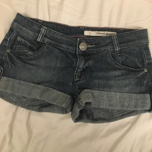 Mörkblå jeansshorts från DKNY - Snygga mörkblå jeansshorts från DKNY med dekorativa knappar och uppvikta ben. Perfekta för en avslappnad stil. Klassisk femficksdesign med broderade detaljer på bakfickorna.