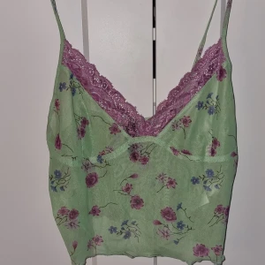 Grön blommig linnetopp från Motel - Söt grön linnetopp från Motel med blommigt mönster i rosa och lila. Toppen har spetsdetaljer vid halsringningen och smala axelband. Perfekt för en somrig look!
