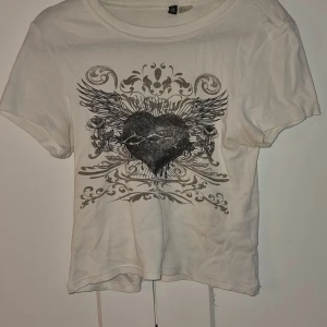 Vit t-shirt med hjärtmotiv från H&M Divided - Säljer en vit t-shirt från H&M Divided med ett stort tryck av ett hjärta med vingar och dekorativa detaljer. T-shirten är kortärmad och har en normal passform. Perfekt för en avslappnad stil.
