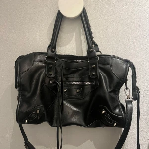 Svart axelväska med dragkedja - Snygg svart axelväska i skinn med dragkedja och dekorativa detaljer. Väskan har både handtag och en justerbar axelrem för flexibel användning.