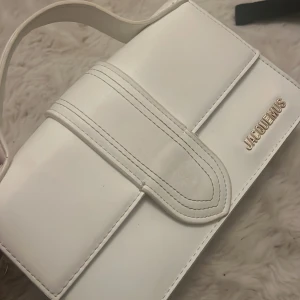 Vit handväska från Jacq - Snygg vit handväska i stilren design. Väskan har ett elegant handtag och märkets logotyp i metall på framsidan. Äkta men smutsig inuti av läppennor så det går självklart att tvätta bort 