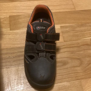 Svarta arbetsskor från Jalas - Svarta arbetsskor från Jalas med orange detaljer och kardborreband för enkel justering. Skorna har en robust design och är certifierade enligt EN ISO 20345:2011 S1 SRC. Skorna har även metall tår så du kan arbeta säkert med skorna på.
