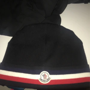 Svart mössa från Moncler - Snygg svart mössa från Moncler med röd, vit och blå randdetalj. Tillverkad i Italien Perfekt för kyliga dagar.    