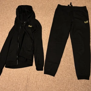 Svart hoodie och byxor från EA7 - Snygg svart hoodie från EA7 med dragkedja och gul logga på bröstet. Perfekt för en avslappnad stil med långa ärmar och huva. Matcha med de svarta byxorna för en komplett look.