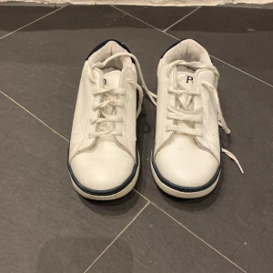 Vita sneakers från Polo - Snygga vita sneakers från Polo med marinblå detaljer och snörning. Perfekta för en avslappnad stil. Märkeslogga på sidan och hälen ger en klassisk touch.