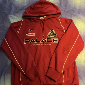 Röd hoodie från Palace - Säljer en snygg röd hoodie från Palace med coola vita detaljer och tryck. Den har en stor logga på framsidan och texten 'Palasonic' som ger en unik touch. Perfekt för höst och vinter med sin mysiga passform och långa ärmar. Huvan är också ett plus för extra värme. Passar perfekt för en casual look eller streetwear-stil.