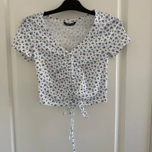 Vit blommig topp med knytning - Säljer en superfin vit topp med blått blommönster. Den har en v-ringning och korta ärmar, perfekt för sommaren. Toppen är croppad och har en snygg knytning framtill som ger en extra detalj.