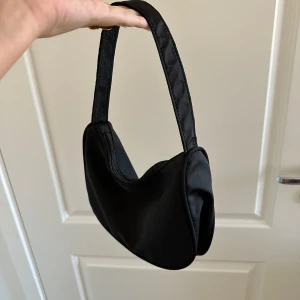 Svart axelväska med dragkedja - Säljer en stilren svart axelväska i syntetmaterial. Väskan har en praktisk dragkedja och en enkel design som passar till alla tillfällen. Perfekt för både vardag och fest!