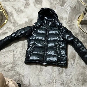 Svart pufferjacka från Moncler - Snygg svart pufferjacka från Moncler med glansig finish och huva. Jackan har en dragkedja framtill och praktiska fickor med knappar. Perfekt för kyliga dagar.