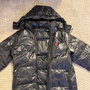Svart dunjacka från Polo Ralph Lauren - Snygg svart dunjacka från Polo Ralph Lauren med huva och dragkedja. Jackan har ett rött broderat emblem på bröstet och praktiska fickor med dragkedjor. Perfekt för kyliga dagar.priset kan diskuterat. Ifall du är intresserad kan skicka mer bilder.