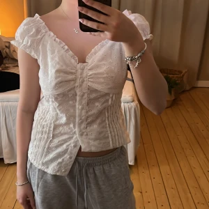 Vit blus med spetsdetaljer - Charmig vit blus med spetsdetaljer och volangärmar. Blusen har en v-ringad design och knappar framtill. Perfekt för en somrig look. Storlek xs/s