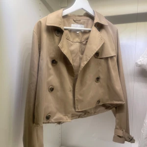 Beige croppad trenchcoat - Snygg beige croppad trenchcoat med knappar och bälte vid ärmsluten. Perfekt för en stilren look under höst och vår. Jackan har en klassisk krage och dubbelknäppning framtill. Den är uppsydd där nere och har lite små fläckar men inget som stör enligt mig men därav priset! Kolla gärna in det andra jag säljer!💘